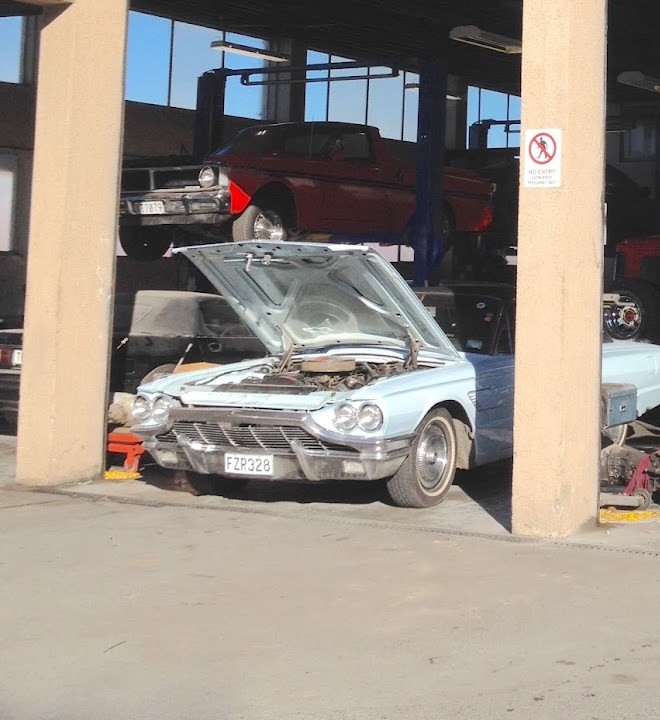 A classic Thunderbird with its bonnet up and a Falcon on the hoist inside the workshop